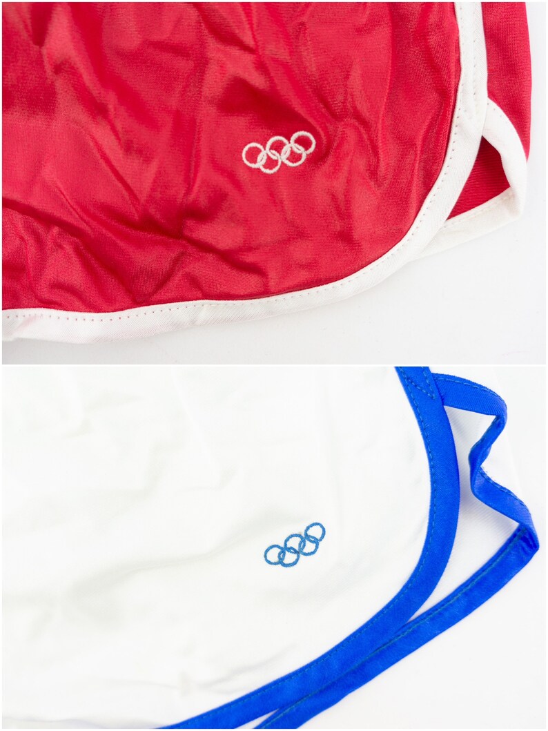 May include: Two pairs of athletic shorts with white and blue trim. One pair is red with a white Olympic rings logo. The other pair is white with a blue Olympic rings logo.
