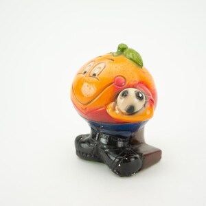 May include: Orange-shaped ceramic figurine of a soccer player holding a soccer ball. The figurine is wearing a red and blue jersey, black shorts, and black shoes.