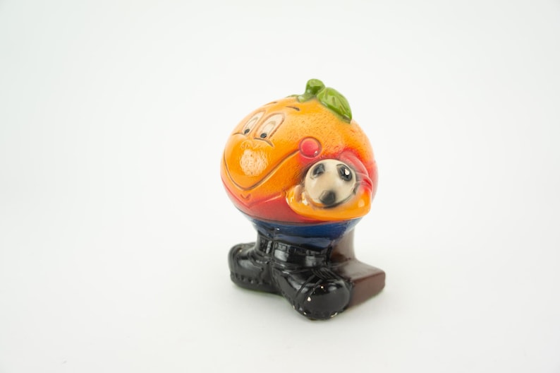 May include: Orange-shaped ceramic figurine of a soccer player holding a soccer ball. The figurine is wearing a red and blue jersey, black shorts, and black shoes.