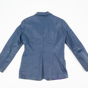 K&ouml;nnte beinhalten: Ein marineblauer Lederblazer mit klassischem Design. Die Jacke hat einen Reverskragen, lange &Auml;rmel und eine taillierte Passform. Die R&uuml;ckseite der Jacke ist sichtbar und zeigt die N&auml;hte und Konstruktionsdetails. Der Blazer liegt flach auf einer wei&szlig;en Oberfl&auml;che.