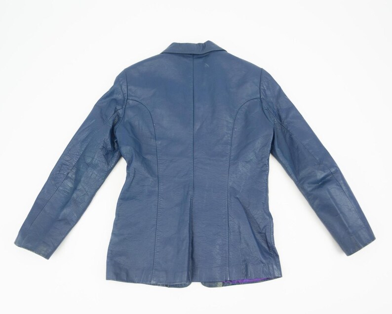 K&ouml;nnte beinhalten: Ein marineblauer Lederblazer mit klassischem Design. Die Jacke hat einen Reverskragen, lange &Auml;rmel und eine taillierte Passform. Die R&uuml;ckseite der Jacke ist sichtbar und zeigt die N&auml;hte und Konstruktionsdetails. Der Blazer liegt flach auf einer wei&szlig;en Oberfl&auml;che.