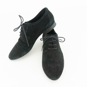 Op de afbeelding: Een paar zwarte su&egrave;de veterschoenen, een met een geperforeerd ontwerp.