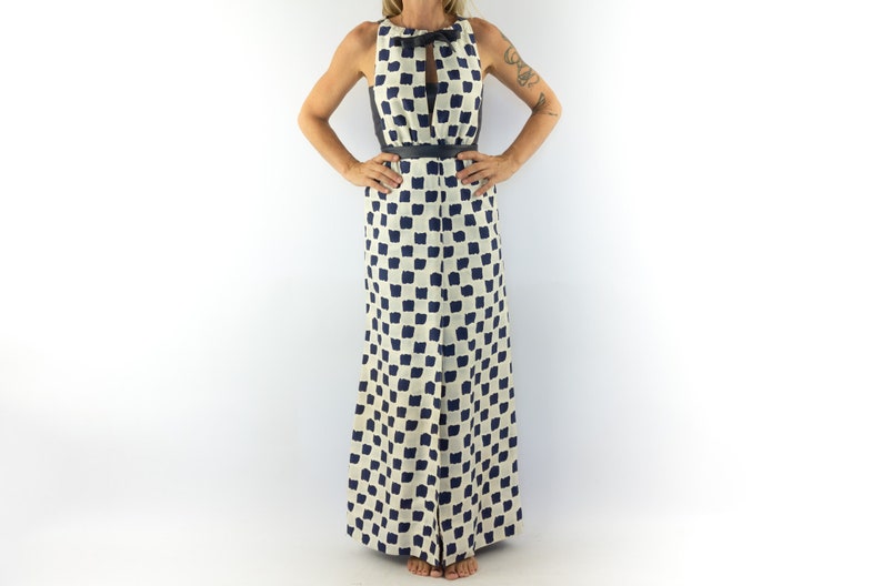 May include: A white maxi dress with a black and white geometric pattern. The dress has a sleeveless design and a keyhole neckline with a bow tie detail. The dress is belted at the waist.