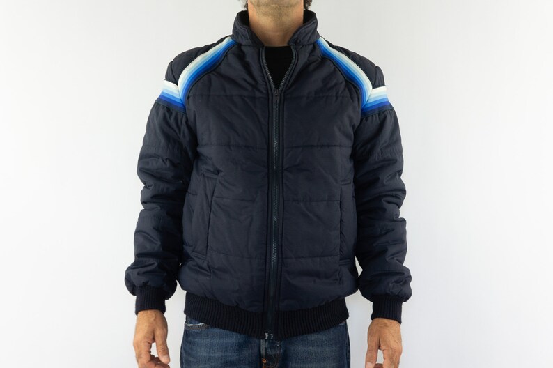 May include: A navy blue puffer jacket with a zipper closure and a ribbed knit collar, cuffs, and waistband. The jacket features a colorful striped design on the shoulders.