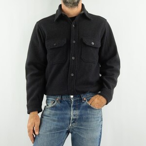 May include: A dark charcoal gray button-down shirt with two chest pockets. The shirt has a classic collar and is made of a thick, textured fabric. The model is wearing blue jeans.