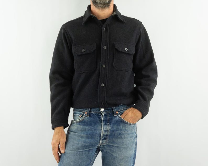 May include: A dark charcoal gray button-down shirt with two chest pockets. The shirt has a classic collar and is made of a thick, textured fabric. The model is wearing blue jeans.