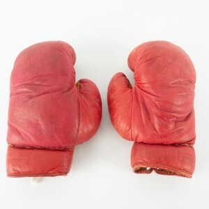 May include: Pair of vintage red boxing gloves. The gloves are made of leather and show signs of wear. The gloves are designed for boxing and other combat sports. The gloves are a vibrant red color.