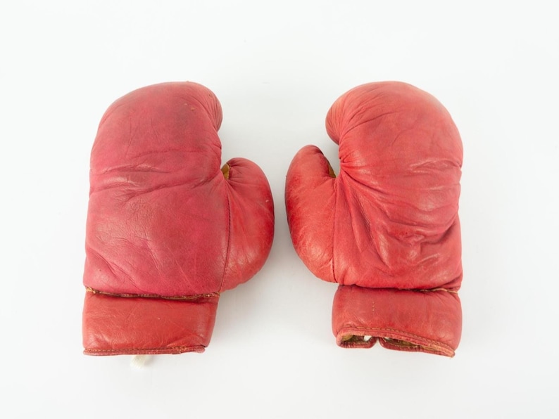 May include: Pair of vintage red boxing gloves. The gloves are made of leather and show signs of wear. The gloves are designed for boxing and other combat sports. The gloves are a vibrant red color.