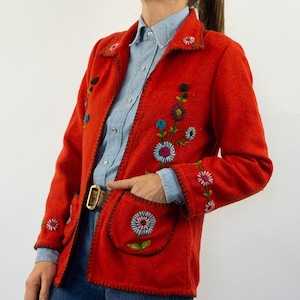 May include: A red jacket with floral embroidery. The jacket has a collar and pockets. The embroidery is in shades of blue, green, pink, and yellow.