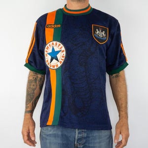 May include: Navy blue Adidas soccer jersey with orange and green accents. Features the Newcastle Brown Ale logo, a star, and the Newcastle United crest. The jersey has orange stripes on the sleeves and a green collar.