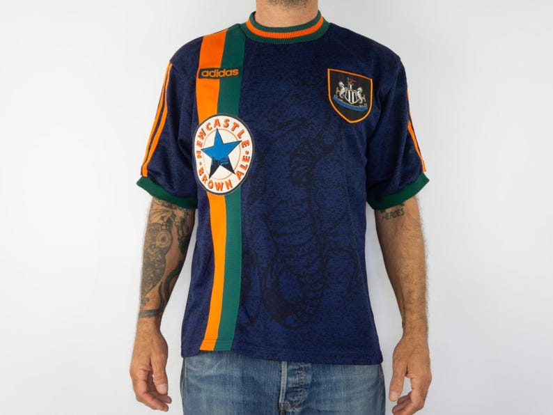 May include: Navy blue Adidas soccer jersey with orange and green accents. Features the Newcastle Brown Ale logo, a star, and the Newcastle United crest. The jersey has orange stripes on the sleeves and a green collar.