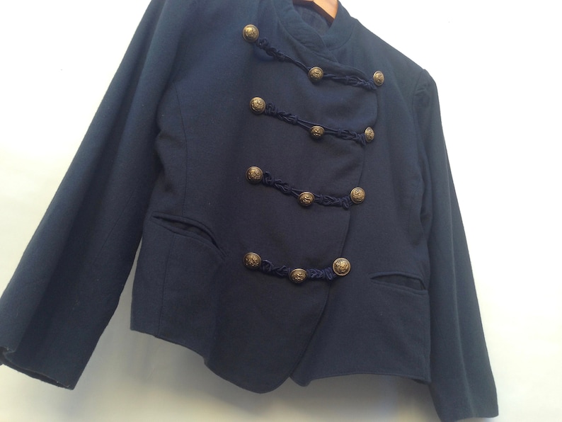 Pode incluir: Um blazer azul escuro com fecho de dupla abotoadura e bot&otilde;es dourados. O blazer tem um design &uacute;nico com guarni&ccedil;&atilde;o tran&ccedil;ada em torno dos bot&otilde;es.