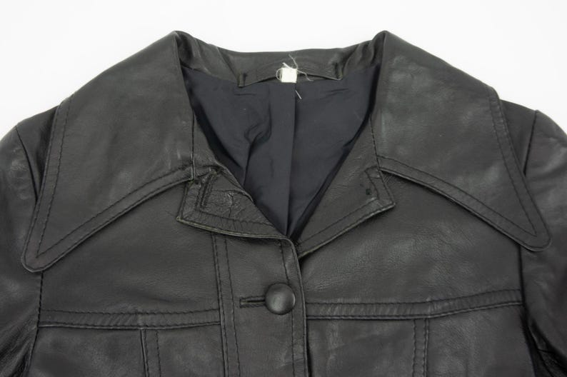 May include: Close-up of a black leather jacket with a large collar and a single button closure. The jacket has a classic design with visible stitching details and a tailored fit. The garment appears to be vintage.