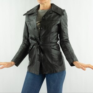 May include: Black leather jacket with a notched collar, button closure, and a tie belt. The jacket has long sleeves and pockets. The jacket is worn over a gray t-shirt and blue jeans.