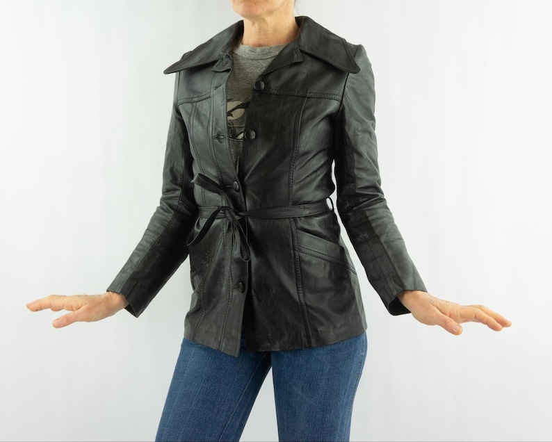 May include: Black leather jacket with a notched collar, button closure, and a tie belt. The jacket has long sleeves and pockets. The jacket is worn over a gray t-shirt and blue jeans.