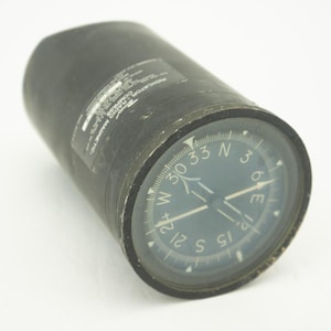 May include: A black cylindrical magnetic compass with a dark blue face. The compass has white markings for direction and degrees. The text "INDICATOR RADIO COMPASS MAGNETIC" is visible on the side.