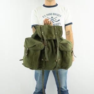 May include: An olive green canvas backpack with multiple pockets and leather straps. The backpack has a flap closure and adjustable straps. The bag is designed for outdoor use and travel.