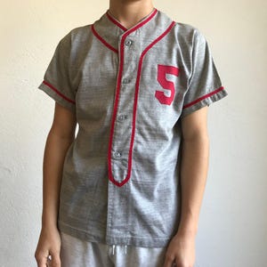 Peut inclure: Un maillot de baseball gris avec des bordures rouges et le numéro 5 sur la poitrine gauche. Le maillot a une fermeture boutonnée sur le devant et des manches courtes. La bordure rouge souligne le col, la patte de boutonnage et les bords des manches.