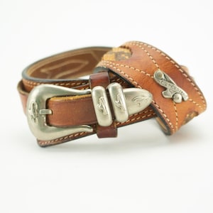 Puede incluir: Un cinturón de cuero marrón con una hebilla plateada y costuras decorativas. La hebilla tiene un diseño floral y el cinturón está estampado con un patrón.