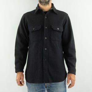 May include: A dark gray wool shirt with a button-down front, two chest pockets, and long sleeves. The shirt has a classic collar and is worn over a black undershirt. The model is wearing blue jeans.