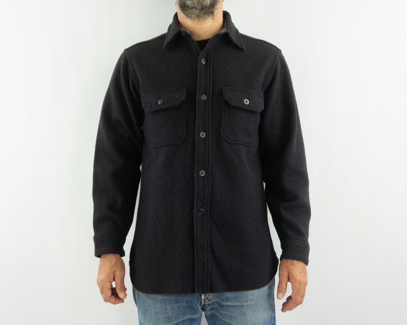 May include: A dark gray wool shirt with a button-down front, two chest pockets, and long sleeves. The shirt has a classic collar and is worn over a black undershirt. The model is wearing blue jeans.