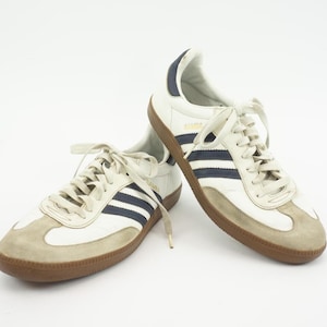 Op de afbeelding: Een paar witte lage sneakers met drie donkerblauwe strepen aan de zijkanten, lichtbruine suède overlays bij de neus en hiel, witte veters en een rubberen zool. Het woord SAMBA staat in gouden letters op de zijkant van elke schoen gedrukt.