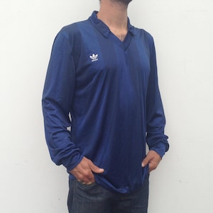 K&ouml;nnte beinhalten: Ein lang&auml;rmeliges marineblaues Poloshirt mit einem dezenten vertikalen Streifenmuster und einem wei&szlig;en Adidas-Trefoil-Logo auf der rechten Brust. Das Shirt hat einen V-Ausschnitt und besteht aus einem glatten, leichten Sportstoff.