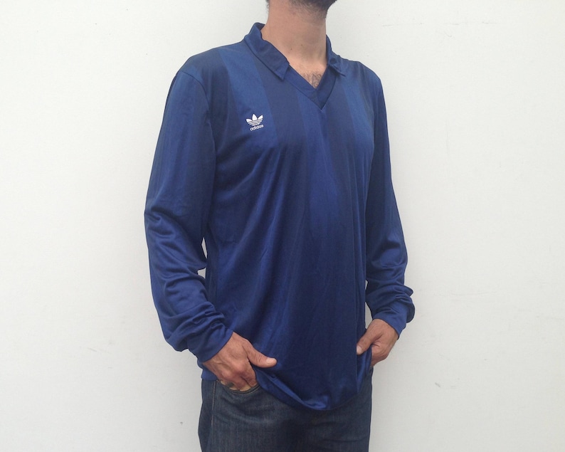 K&ouml;nnte beinhalten: Ein lang&auml;rmeliges marineblaues Poloshirt mit einem dezenten vertikalen Streifenmuster und einem wei&szlig;en Adidas-Trefoil-Logo auf der rechten Brust. Das Shirt hat einen V-Ausschnitt und besteht aus einem glatten, leichten Sportstoff.