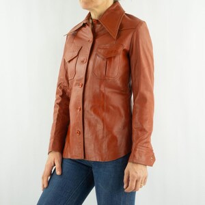 May include: A vintage, rust-colored leather shirt with a pointed collar, two chest pockets, and button closures. The long-sleeved shirt is worn with blue jeans. The leather appears to be in good condition.