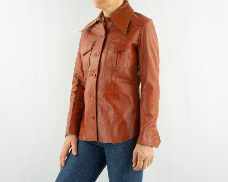 May include: A vintage, rust-colored leather shirt with a pointed collar, two chest pockets, and button closures. The long-sleeved shirt is worn with blue jeans. The leather appears to be in good condition.