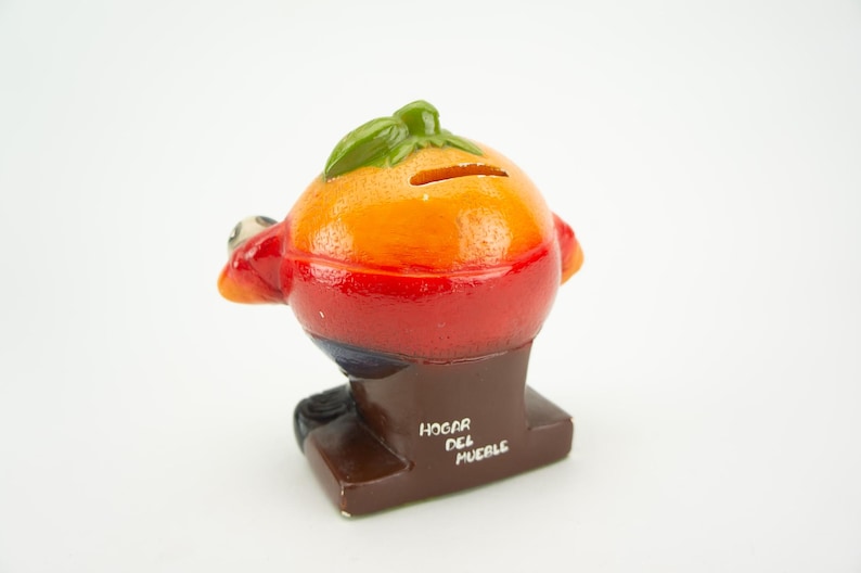 May include: A ceramic piggy bank shaped like a cartoon character with a red, orange, and yellow body. The piggy bank has a green leaf on top and a slot for coins on the back. The base of the piggy bank is brown and has the text "HOGAR DEL MUEBLE" written on it.