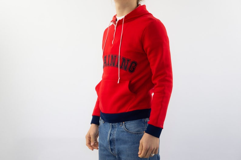 K&ouml;nnte beinhalten: Roter Kapuzenpullover mit einem dunkelblauen "TRAINING"-Logo auf der Vorderseite. Der Pullover hat einen halben Rei&szlig;verschluss und einen dunkelblauen Bund am unteren Rand.