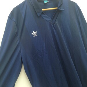 K&ouml;nnte beinhalten: Ein lang&auml;rmeliges marineblaues Hemd mit V-Ausschnitt und einem dezenten vertikalen Streifenmuster. Das Hemd zeigt ein wei&szlig;es Adidas-Trefoil-Logo auf der rechten Brust. Es wird auf einem Holzkleiderb&uuml;gel vor wei&szlig;em Hintergrund pr&auml;sentiert.