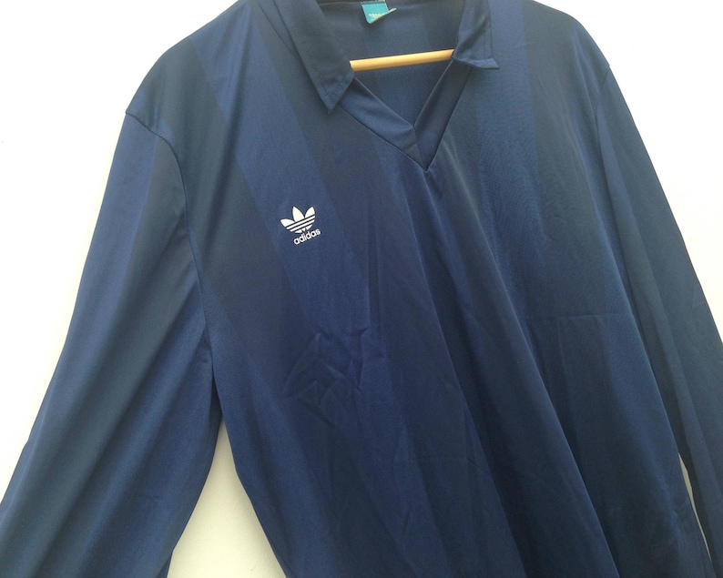 K&ouml;nnte beinhalten: Ein lang&auml;rmeliges marineblaues Hemd mit V-Ausschnitt und einem dezenten vertikalen Streifenmuster. Das Hemd zeigt ein wei&szlig;es Adidas-Trefoil-Logo auf der rechten Brust. Es wird auf einem Holzkleiderb&uuml;gel vor wei&szlig;em Hintergrund pr&auml;sentiert.