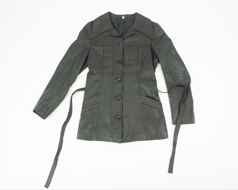 May include: A black leather jacket with a button-up front, long sleeves, and a tie belt. The jacket has a collared neckline and two front pockets. The jacket is displayed on a white background.