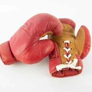 May include: A pair of vintage boxing gloves. One glove is red, the other is brown and red with white laces. The gloves are made of leather and show signs of wear.