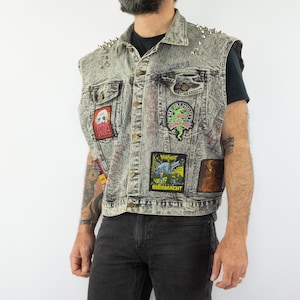Peut inclure: Un gilet en jean gris avec des clous argentés et divers patchs. Les patchs représentent un crâne, un monstre, un logo de groupe et un design graphique. Le gilet a un aspect vieilli.