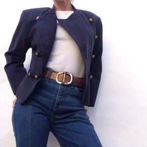 Pode incluir: Uma jaqueta azul marinho cropped com bot&otilde;es dourados e uma camiseta branca por baixo. A jaqueta tem fecho de dupla abotoadura e uma silhueta ligeiramente ajustada.