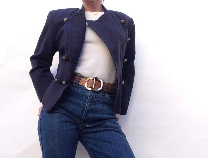 Pode incluir: Uma jaqueta azul marinho cropped com bot&otilde;es dourados e uma camiseta branca por baixo. A jaqueta tem fecho de dupla abotoadura e uma silhueta ligeiramente ajustada.