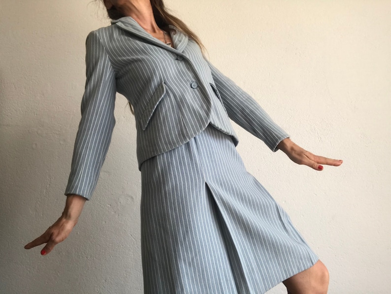 Puede incluir: Un traje azul claro de rayas finas con una chaqueta y una falda. La chaqueta tiene un cierre de un solo bot&oacute;n y la falda tiene un frente plisado.