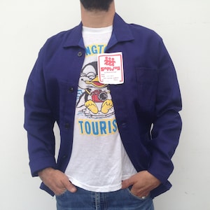 May include: A blue work jacket with a white t-shirt underneath. The t-shirt has a cartoon penguin wearing a camera and the text "TOURIST" on it. A small red and white sticker with the text "SAPLING" is attached to the jacket.
