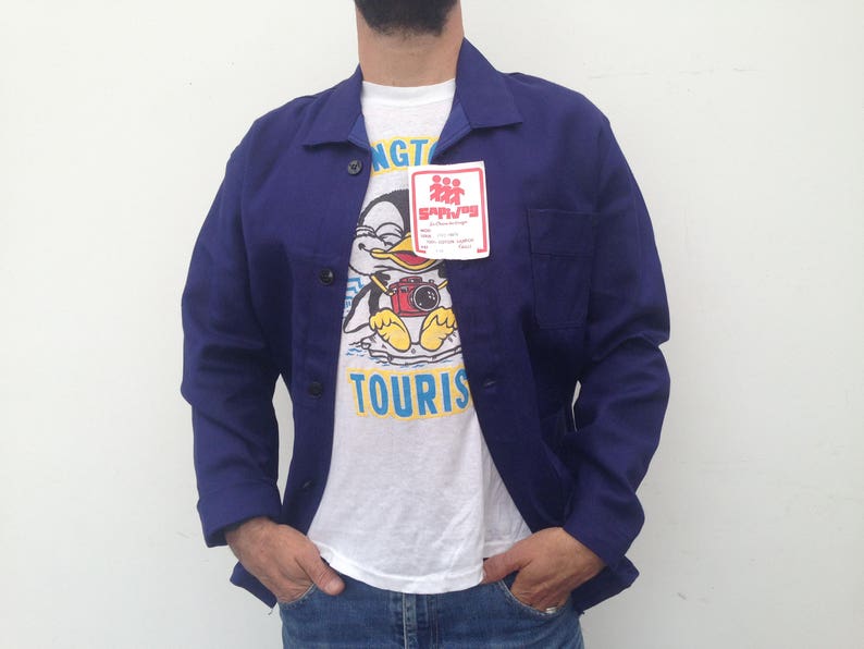 May include: A blue work jacket with a white t-shirt underneath. The t-shirt has a cartoon penguin wearing a camera and the text "TOURIST" on it. A small red and white sticker with the text "SAPLING" is attached to the jacket.
