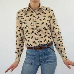 May include: A beige button-down shirt with a black and white print of running lions. The shirt has long sleeves and a collared neckline.