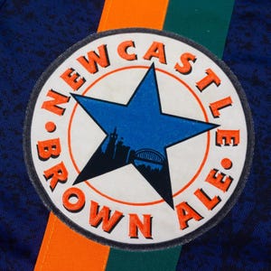 May include: Close-up of a navy blue fabric with a circular patch. The patch features a blue star with a city silhouette, surrounded by the words "NEWCASTLE BROWN ALE" in orange. Orange and green stripes are visible.
