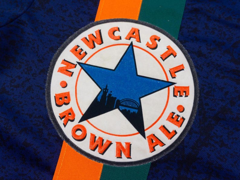May include: Close-up of a navy blue fabric with a circular patch. The patch features a blue star with a city silhouette, surrounded by the words "NEWCASTLE BROWN ALE" in orange. Orange and green stripes are visible.