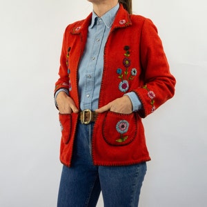 May include: A red jacket with embroidered flowers in shades of blue, green, and pink. The jacket has a collar and pockets. The model is wearing a light blue button-down shirt and blue jeans.