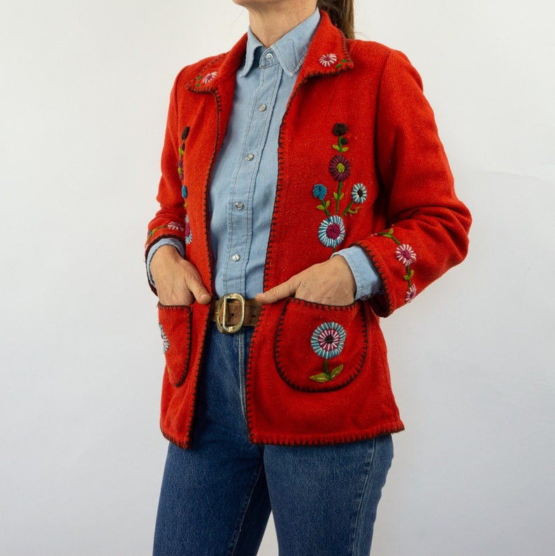 May include: A red jacket with embroidered flowers in shades of blue, green, and pink. The jacket has a collar and pockets. The model is wearing a light blue button-down shirt and blue jeans.