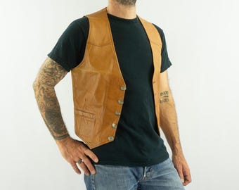 May include: A tan leather vest with a classic design. The vest has a V-neck, a button-up front, and two front pockets. The vest is worn over a black t-shirt and blue jeans. The vest appears to be vintage.