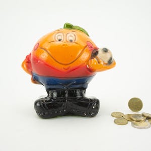 May include: Orange ceramic piggy bank shaped like a cartoon character wearing a red, blue, and white soccer uniform. The character is holding a soccer ball and has a smiling face. There are several gold coins scattered on the white surface.