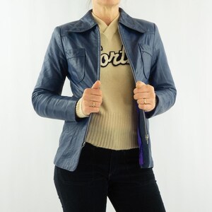 K&ouml;nnte beinhalten: Blaue Lederjacke mit Kragen und Rei&szlig;verschluss. Die Jacke hat zwei Vordertaschen und wird &uuml;ber einem beigen Pullover getragen. Die Person tr&auml;gt auch eine schwarze Hose.